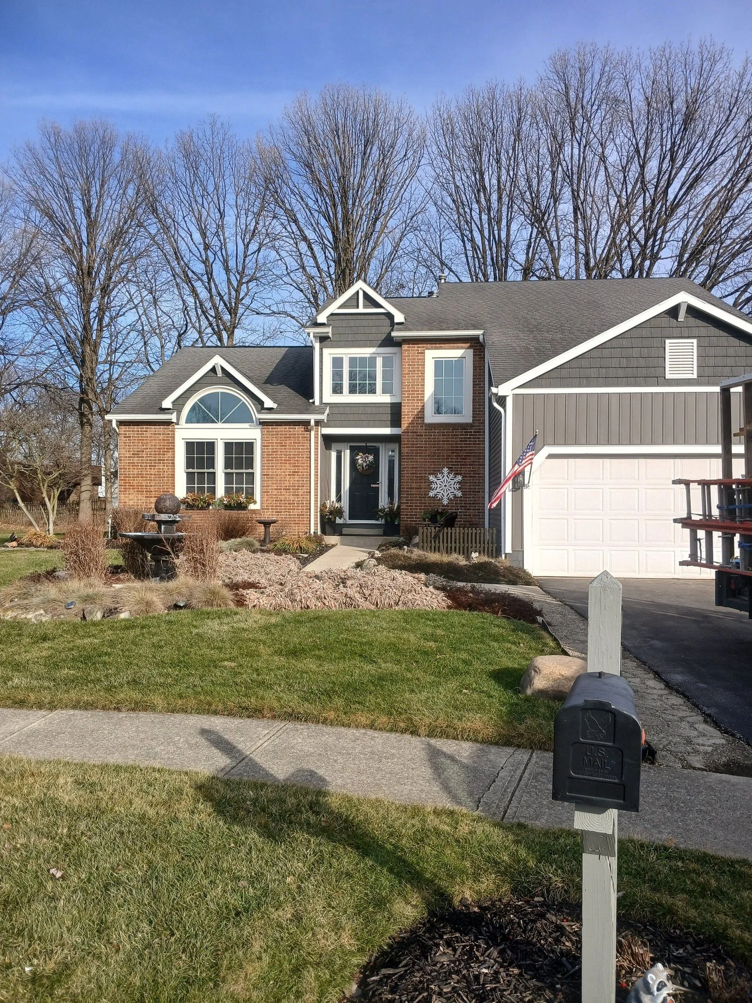 Suburban brick house with double garage, front porch with a wreath, manicured lawn, and American flag by the entrance.