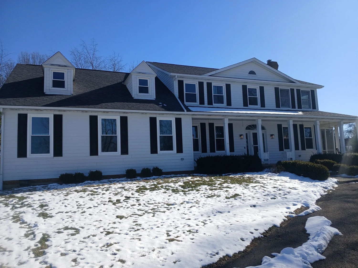 White two-story house with dark shutters, porch, and dormer windows, surrounded by snowy lawn.