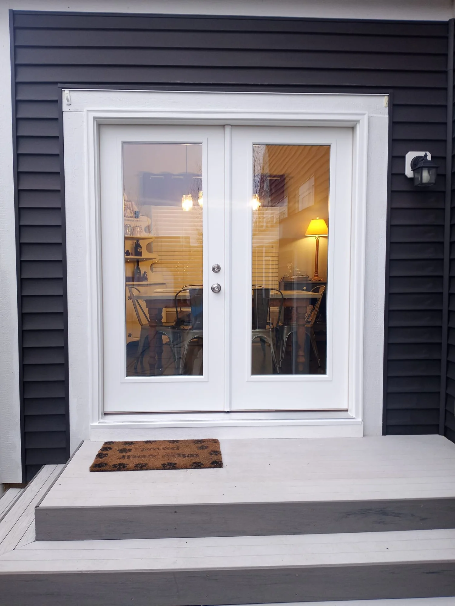 Exterior view of white double glass doors on a building with a dark grey facade, small steps leading up, a welcome mat, and interior lights visible through the doors.