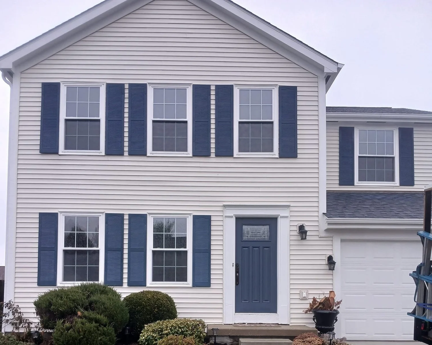 Two-story house with beige siding, blue shutters and door, garage, and front shrubs.
