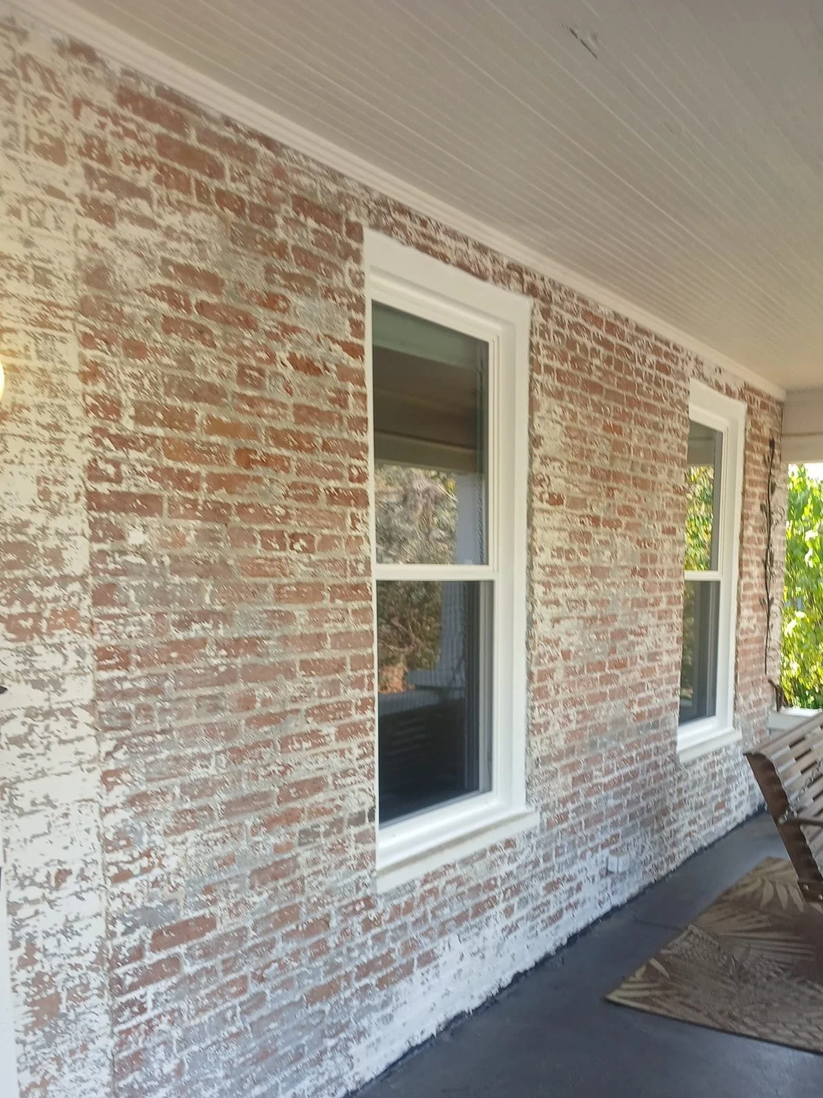 Red brick wall with white distressed paint, two windows, and a wooden bench on a porch